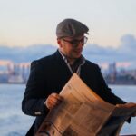 Casually dressed man reading a newspaper on a boat with city skyline backdrop at sunset.