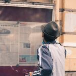 A man in a hat reads a newspaper on an outdoor message board.