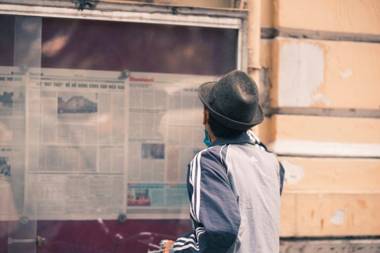 A man in a hat reads a newspaper on an outdoor message board.