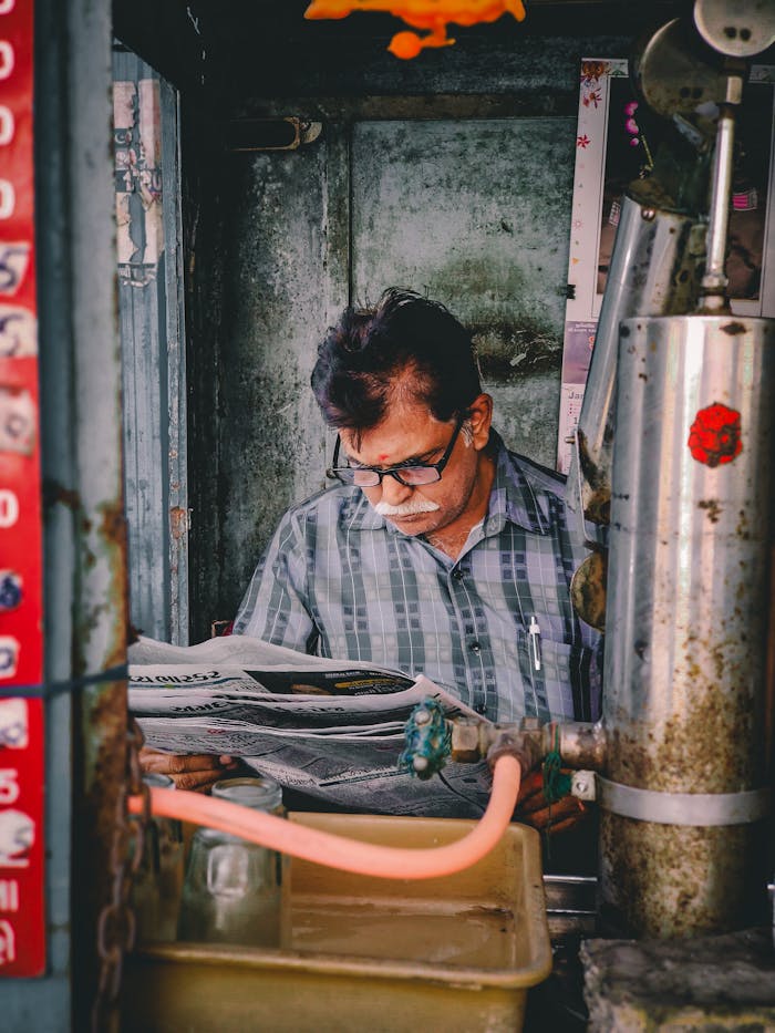 South Asian man reading a newspaper in an Ahmedabad street booth, surrounded by vending equipment.