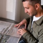 A man in a casual jacket reads a newspaper at a table indoors.