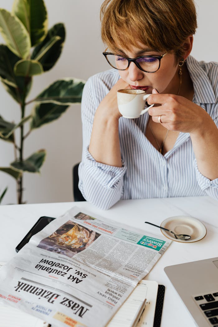 Crop concentrated female in eyeglasses and casual clothes sitting at table and leaning on hand while enjoying cup of hot coffee and reading newspaper near green plant