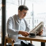 Adult man reading a newspaper while holding a coffee cup in a cozy cafe setting.