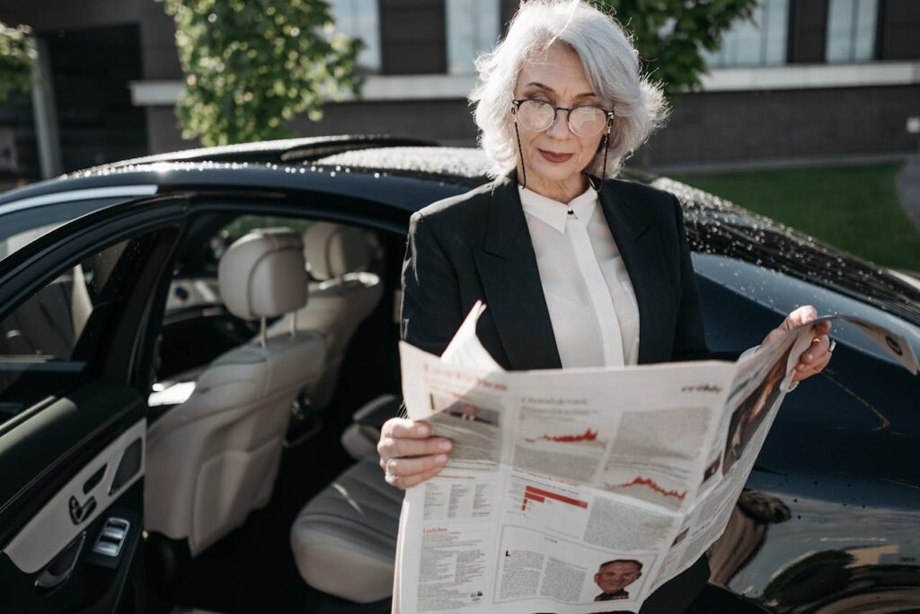 Elegant senior woman in formal attire reading a newspaper next to a luxury car outdoors.