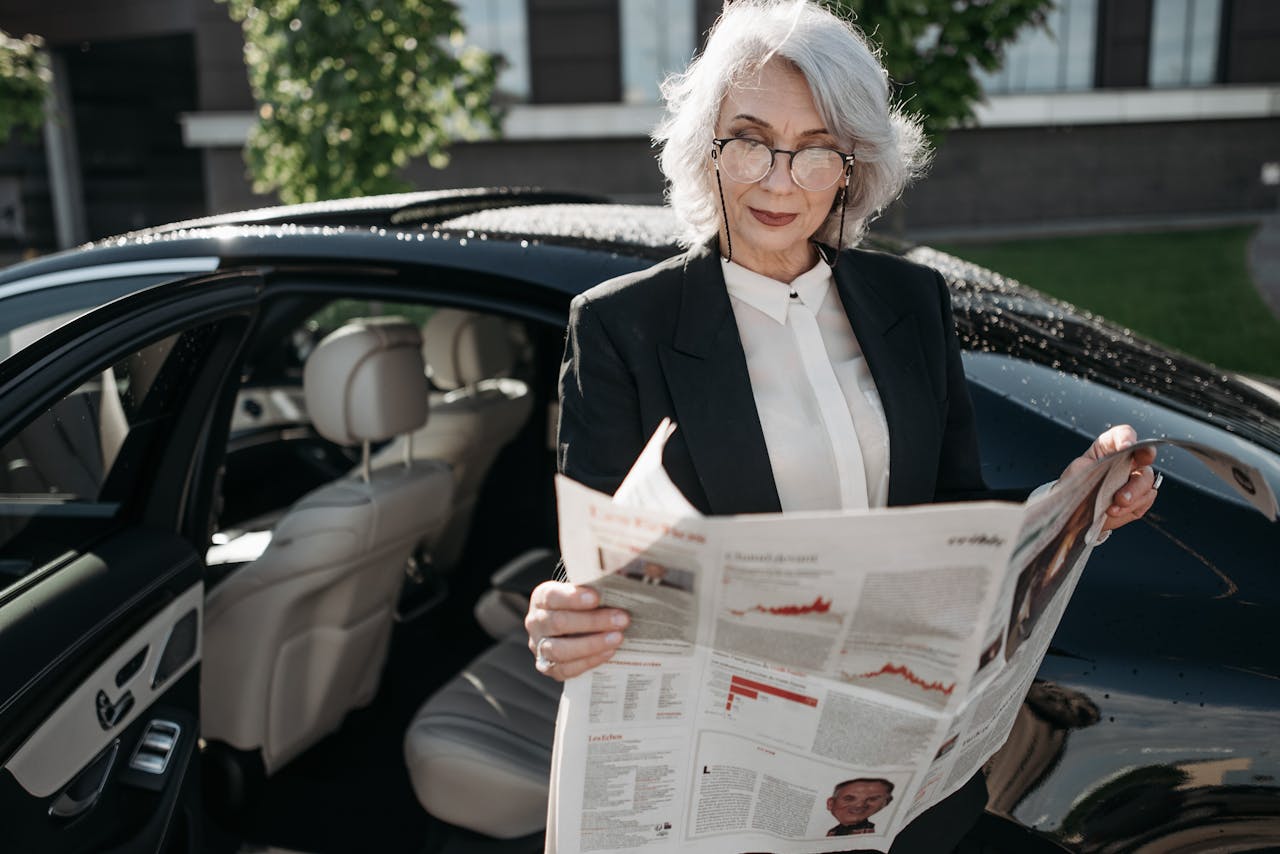 Elegant senior woman in formal attire reading a newspaper next to a luxury car outdoors.
